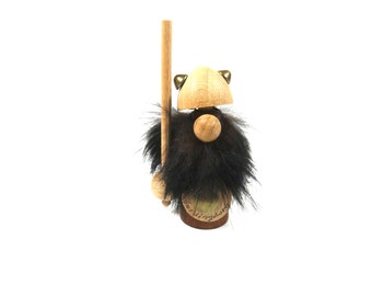 May include: Wooden figurine of a gnome with a black fur beard, wearing a brown hat with gold accents and holding a wooden stick. The gnome is standing on a wooden base with the text "The Kingdom" written on it.