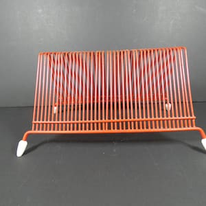 May include: A red metal record holder with a rectangular shape. The holder has vertical bars and white feet. It is designed to hold vinyl records and is a vintage item.
