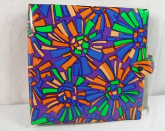 May include: A purple binder with a colorful geometric pattern in shades of orange, blue, green, and white. The binder has a flap with a button closure.