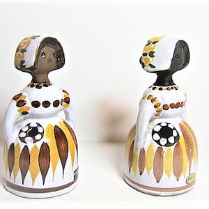 Puede incluir: Dos figuritas de cerámica de mujeres que llevan vestidos blancos con motivos florales marrones y amarillos. Las mujeres tienen caras negras y cubiertas para la cabeza blancas.