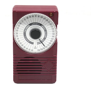 Può includere: Un metronomo rosso in stile vintage con un quadrante bianco e una manopola nera. Il quadrante ha un quadrante con numeri da 40 a 208, e un quadrante più piccolo con numeri da 60 a 184. Il testo "ANDANTE MADE IN W. GERMANY" è stampato sul quadrante. Il metronomo ha un logo a forma di diamante nella parte inferiore.