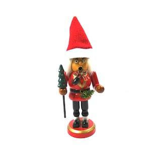Könnte beinhalten: Eine hölzerne Weihnachtsfigur einer Person, die einen roten Weihnachtsmann-Hut mit weißem Besatz und ein rot-schwarzes Outfit trägt und einen kleinen grünen Weihnachtsbaum hält.