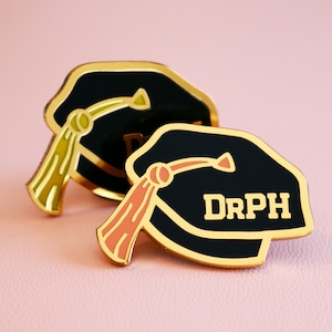 May include: Two gold graduation cap pins. One pin has a green tassel and the other has a pink tassel. The pin with the pink tassel has the text "DRPH" on it.