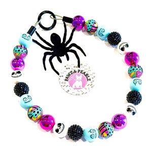 May include: A circular beaded accessory featuring a black spider charm and a round tag with the text PAWZ & PEARLZ. The beads include shiny purple spheres, black textured balls, and light blue beads with printed character faces and patterns.