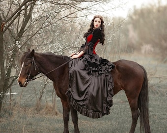 May include: A woman in a black and red dress rides a brown horse in a field with a tree in the background.