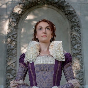 May include: A woman in a purple and gold brocade dress with a large white lace collar. She is standing in front of a stone archway and looking up.