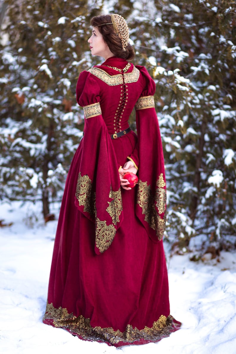 May include: A woman wearing a red velvet gown with gold embroidery and a gold crown stands in a snowy woodland holding a red apple.