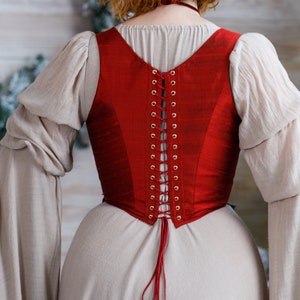 May include: A red corset with a lace-up back and gold eyelets. The corset is worn over a white dress.