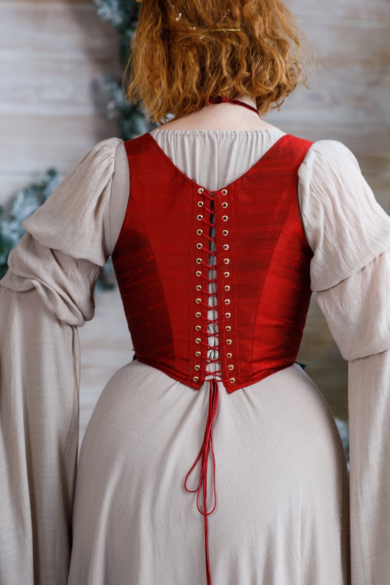May include: A red corset with a lace-up back and gold eyelets. The corset is worn over a white dress.