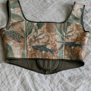 May include: A brown and green corset with a floral and fish print. The corset has a dark green trim and a hook and eye closure at the back.