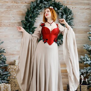 May include: A woman wearing a red corset with floral embellishments over a beige gown with long sleeves. She is standing in front of a wooden wall with a Christmas wreath and a Christmas tree behind her.