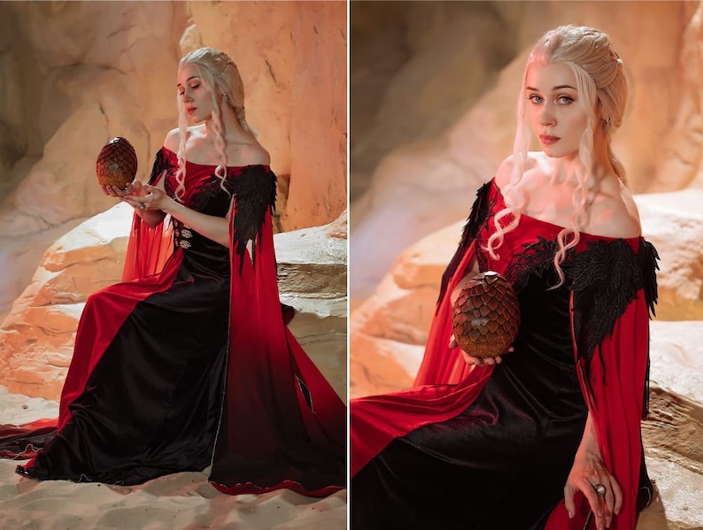 May include: A woman with long, white hair is wearing a red and black dress with a black feathered cape. She is holding a dragon egg in her hands.