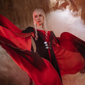 May include: A woman with long white hair wearing a black and red dress with a long flowing cape. The cape is red and black and is flowing in the wind. The woman is standing in front of a rocky background with smoke in the air.
