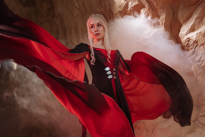 May include: A woman with long white hair wearing a black and red dress with a long flowing cape. The cape is red and black and is flowing in the wind. The woman is standing in front of a rocky background with smoke in the air.