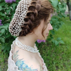 May include: A woman with long brown hair styled in a braid wears a white pearl hair net. She is wearing a pearl necklace and a light brown off-the-shoulder top.