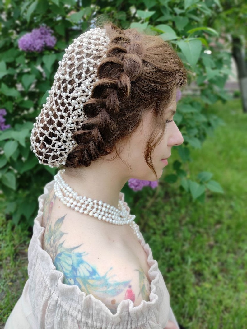 May include: A woman with long brown hair styled in a braid wears a white pearl hair net. She is wearing a pearl necklace and a light brown off-the-shoulder top.