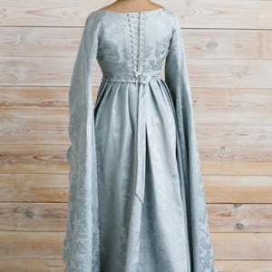 Op de afbeelding: Een lichtblauwe jurk met lange mouwen en een getailleerd lijfje. De jurk heeft een gespeld rugpand en een riem in de taille. De jurk is op een mannequin met een bloemenhoed.