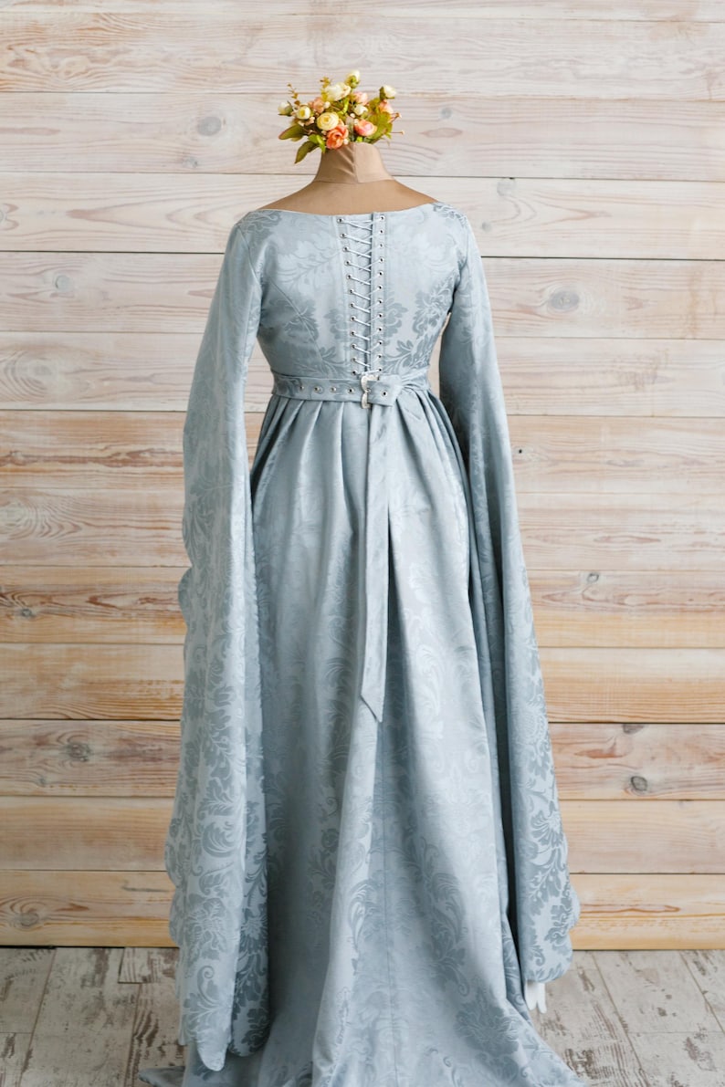 Op de afbeelding: Een lichtblauwe jurk met lange mouwen en een getailleerd lijfje. De jurk heeft een gespeld rugpand en een riem in de taille. De jurk is op een mannequin met een bloemenhoed.