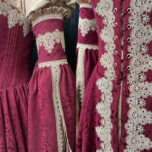 Op de afbeelding: Een diepbordeauxrode jurk met een gestructureerd damastpatroon, versierd met ingewikkelde cr&egrave;mekleurige kanten randen langs het lijfje en de mouwen. Het kledingstuk heeft een vetersluiting aan de voorzijde met goudkleurige oogjes.