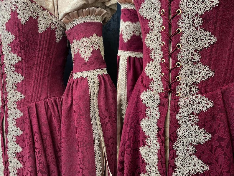 Op de afbeelding: Een diepbordeauxrode jurk met een gestructureerd damastpatroon, versierd met ingewikkelde cr&egrave;mekleurige kanten randen langs het lijfje en de mouwen. Het kledingstuk heeft een vetersluiting aan de voorzijde met goudkleurige oogjes.