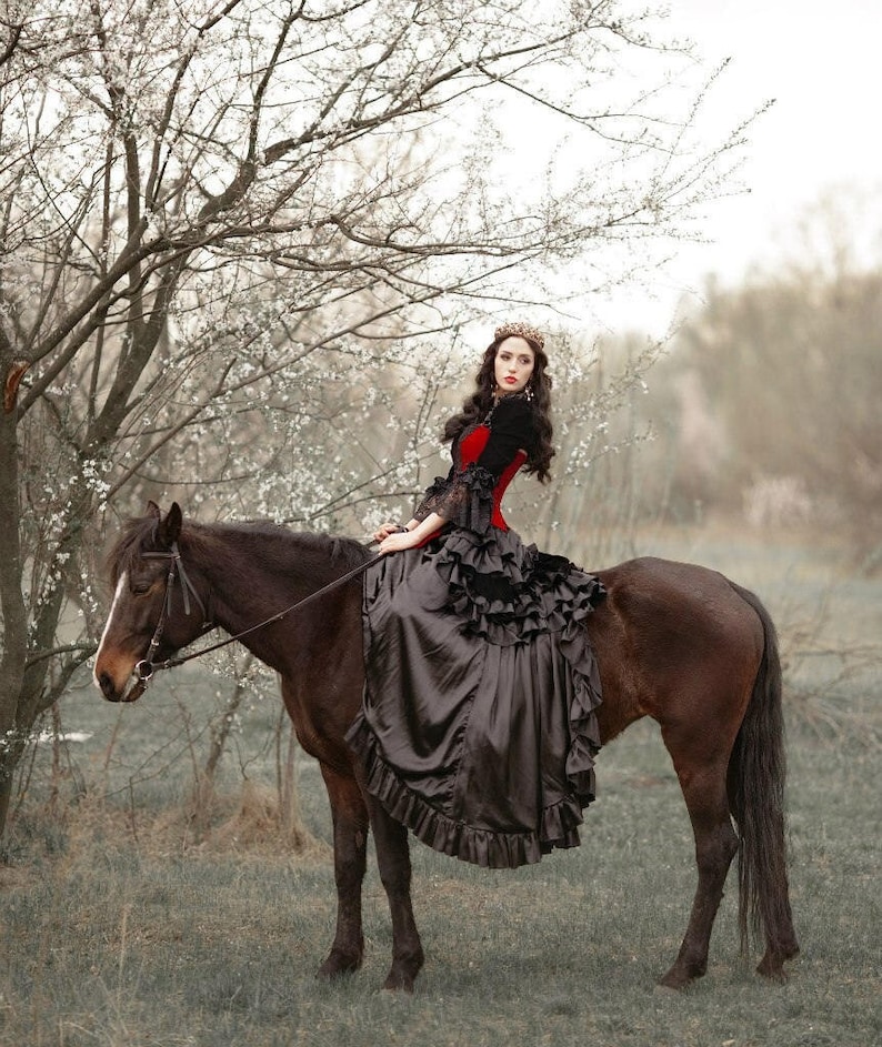 May include: A woman in a black and red dress rides a brown horse in a field with a tree in the background.