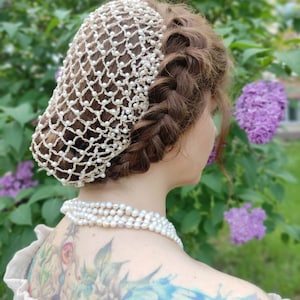 May include: A white crocheted hair net with pearl beads, worn by a person with brown hair. The hair net is worn over a braided hairstyle. The person is wearing a pearl necklace.