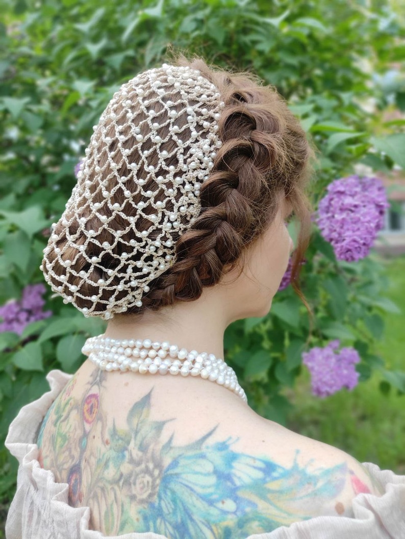 May include: A white crocheted hair net with pearl beads, worn by a person with brown hair. The hair net is worn over a braided hairstyle. The person is wearing a pearl necklace.