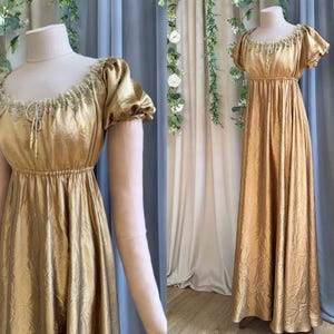 Op de afbeelding: Een vloerlange goudkleurige jurk met een metallic glans, voorzien van korte pofmouwen, een empire-taille en een geplooide ronde halslijn versierd met delicaat kant en een centraal trekkoord met twee bungelende goudkleurige kwastjes.