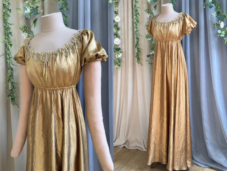 Op de afbeelding: Een vloerlange goudkleurige jurk met een metallic glans, voorzien van korte pofmouwen, een empire-taille en een geplooide ronde halslijn versierd met delicaat kant en een centraal trekkoord met twee bungelende goudkleurige kwastjes.