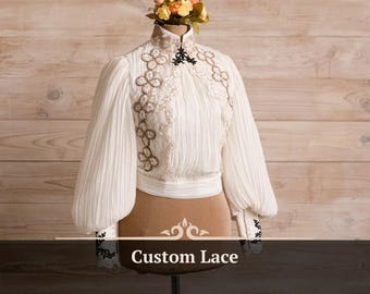 Puede incluir: Blusa blanca de manga larga y cuello alto. La blusa tiene un delicado patrón de encaje con bordados negros y una etiqueta de texto "Custom Lace".
