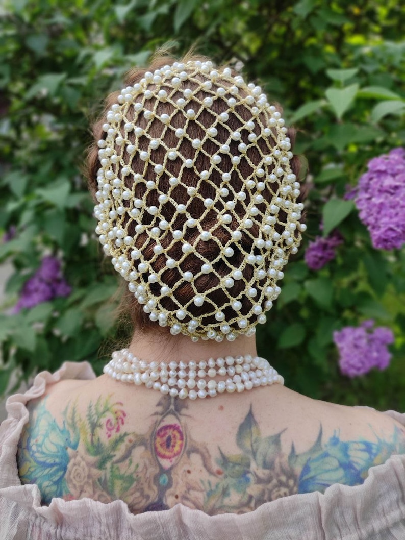 May include: A white pearl hair net with a crocheted design. The net is worn on a person's head and covers the hair. The net is made of small white pearls that are connected by a thin gold thread.
