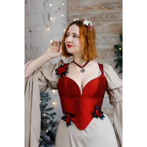 May include: A woman wearing a red corset with floral embellishments and a beige dress with long sleeves. She is wearing a silver necklace with a pendant.