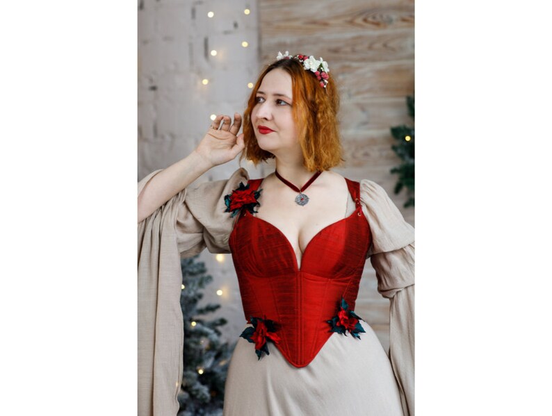 May include: A woman wearing a red corset with floral embellishments and a beige dress with long sleeves. She is wearing a silver necklace with a pendant.
