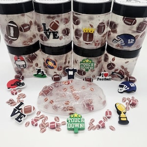 May include: A collection of clear jars filled with a clear gel and football-themed decorations. The jars are stacked, and various football-themed charms and decorations are scattered around, including helmets, footballs, and the words "LOVE" and "TOUCH DOWN!"