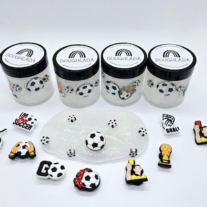May include: Four clear jars with black lids, labeled "Doughlada," filled with glitter and soccer ball decorations. A white, glittery slime with soccer ball charms and various soccer-themed charms are also present.