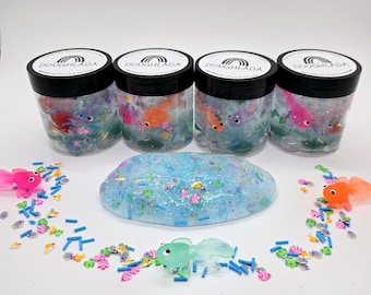 May include: Four clear jars of blue slime with glitter and small plastic fish toys. The slime is spread out on a white surface with more plastic fish toys and sprinkles.