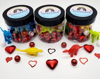 May include: Three clear jars filled with red hearts, white and colored plastic dinosaurs, and colorful beads. The jars have black lids with a rainbow and the text "PLAYDOUGH SENSORY KITS DOUGHLADA" on them.