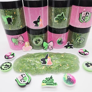 May include: A collection of glittery green slime and pink containers with black lids. The containers have decorative stickers, including a witch silhouette, bows, and a school crest. Additional charms are scattered around the slime.