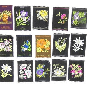 May include: A collection of 16 vintage fabric patches featuring various floral designs. The patches are black with colorful embroidered flowers, including lilies, irises, and roses. The patches are labeled with the word "EUROPA" and the names of the flowers in German.