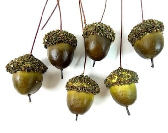 May include: Six green acorn decorations with brown stems and a speckled texture. The acorns are hanging against a white background.