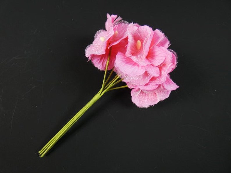 May include: A bouquet of pink silk flowers with green stems. The flowers are arranged in a cluster and have a delicate, ruffled texture.