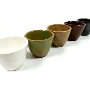 May include: Five ceramic cups in different colours, including white, green, brown, and black. The cups are arranged in a row from left to right, with the white cup at the front and the black cup at the back.