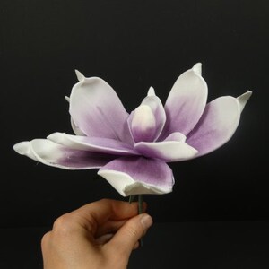 May include: A single artificial magnolia flower with white petals and purple accents. The flower is on a green stem.