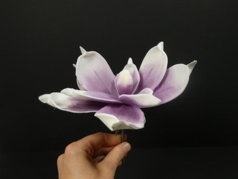 May include: A single artificial magnolia flower with white petals and purple accents. The flower is on a green stem.