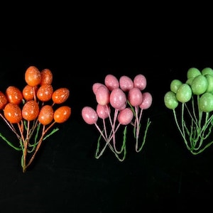 May include: Three clusters of decorative Easter egg stems. One cluster is orange with glitter, another is pink with glitter, and the last is green with glitter. Each stem has a wire base and is set against a black background.