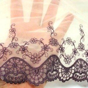 May include: A sheer white fabric with a delicate purple embroidered floral pattern along the bottom edge. The fabric has a scalloped edge and a soft, flowing texture.