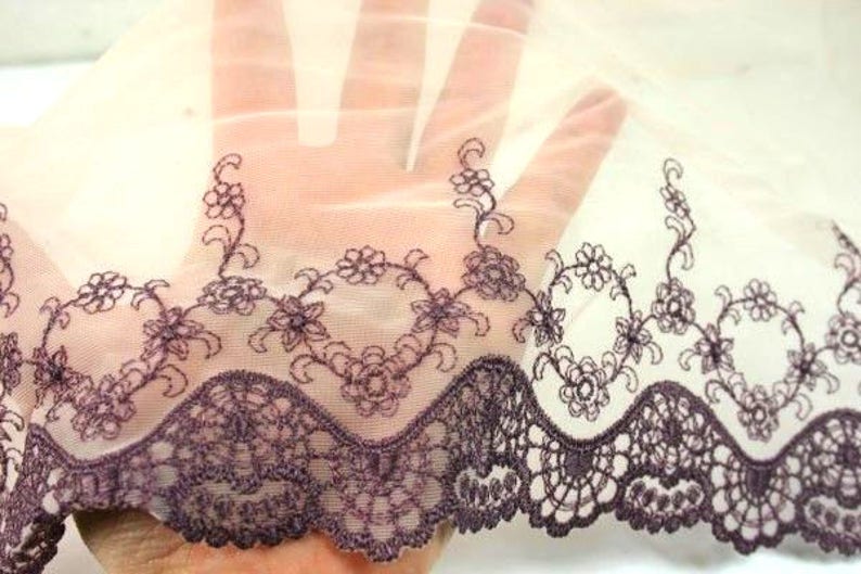 May include: A sheer white fabric with a delicate purple embroidered floral pattern along the bottom edge. The fabric has a scalloped edge and a soft, flowing texture.