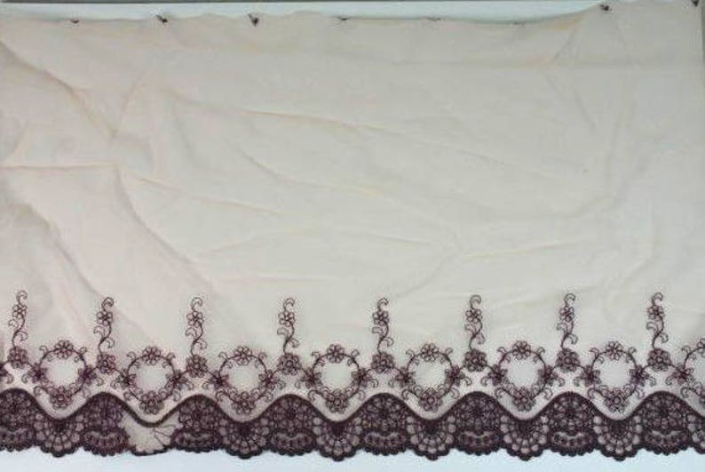 May include: A close-up of a beige sheer fabric with a brown embroidered lace trim. The lace features a scalloped edge and a delicate floral design.