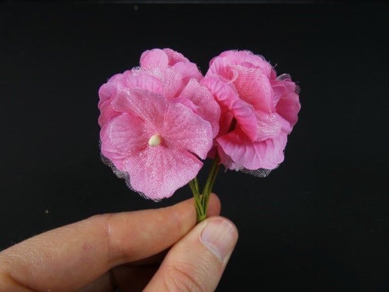 May include: A cluster of pink silk flowers with a delicate, ruffled texture. The flowers are arranged on a stem and have a soft, feminine appearance.
