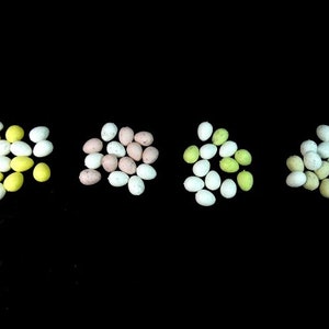 May include: Assortment of small, speckled Easter eggs in yellow, white, pink, and green colours. The eggs are arranged in four groups on a black background.